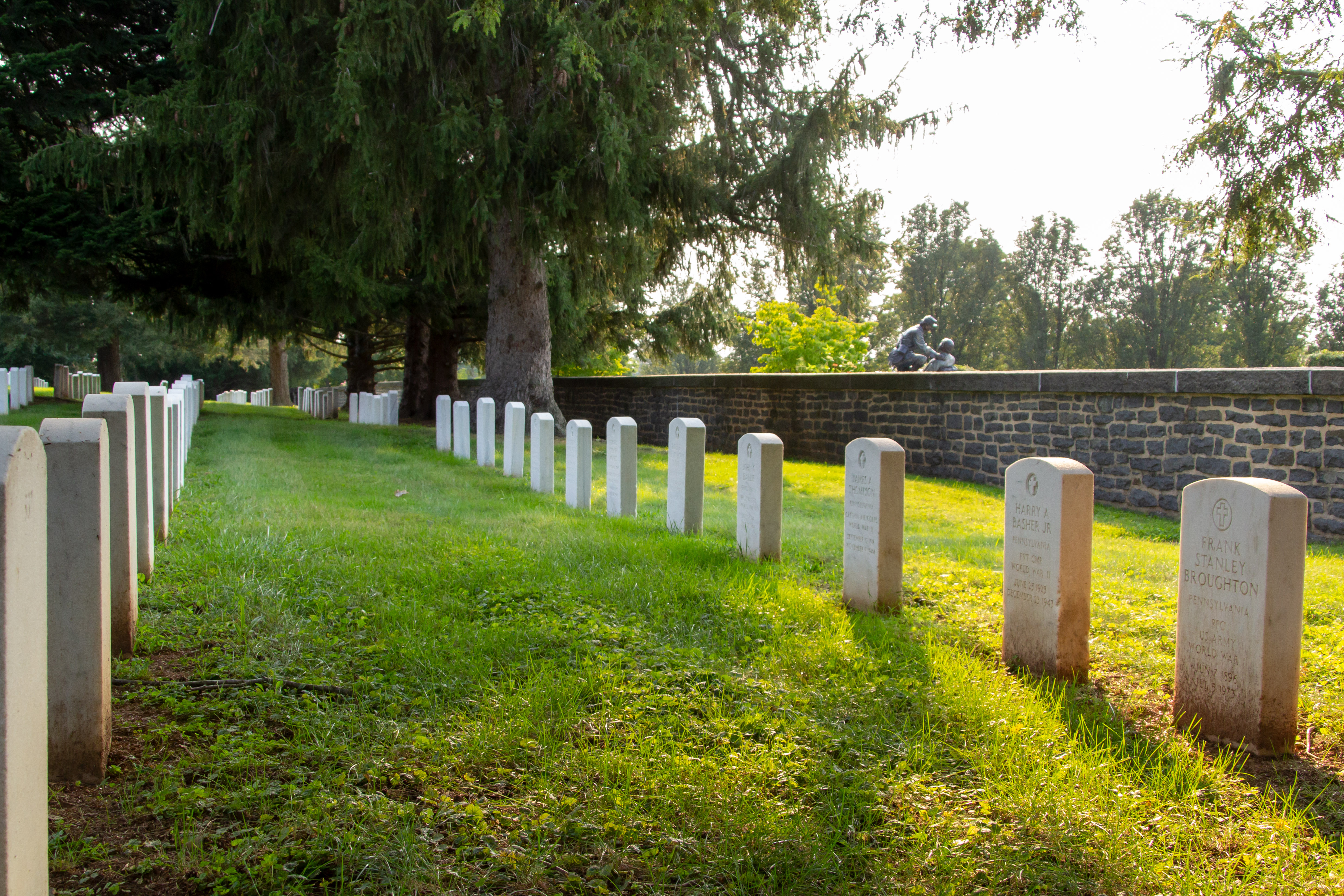 Two rows of white marble gravestones form parallel lines along a dark stone wall in a cemetery. Many mature trees are in the distance.