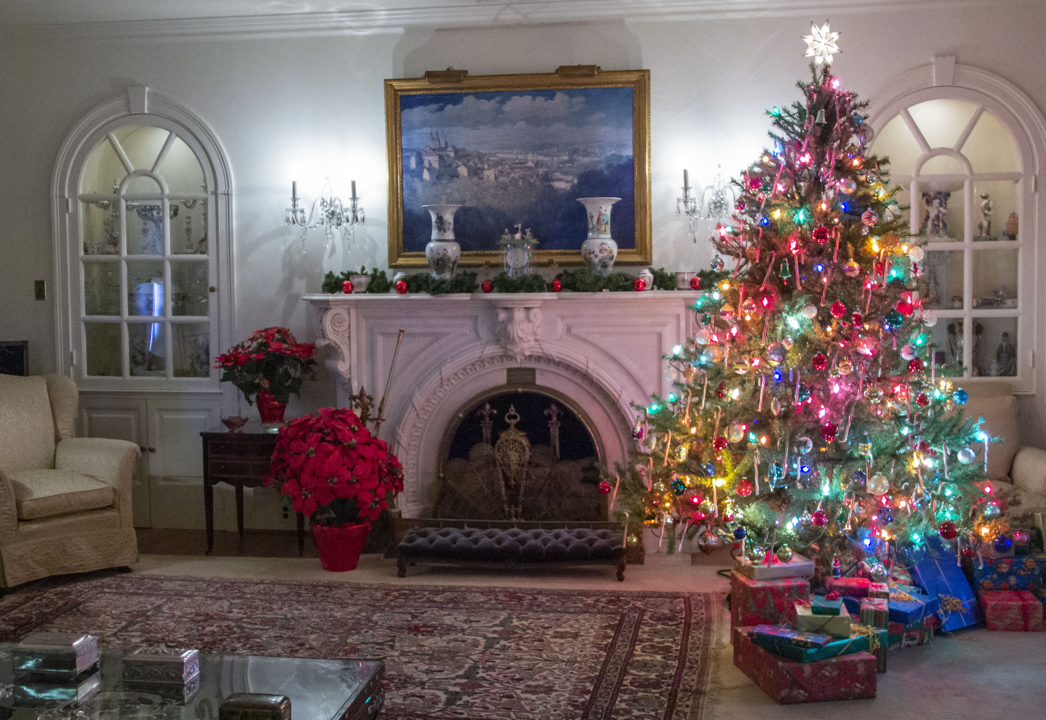 A formal living room is decorated for Christmas. A tall tree stands on the right decorated with colored lights and decorations, a white marble fireplace mantle is in the center with a painting above it, and a poinsettia sits on the floor to the left.