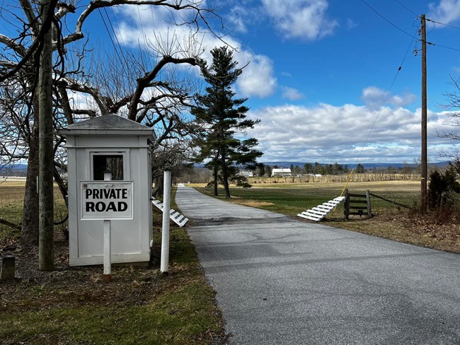 A white pained sign saying Private Road sits next to a white guard shack and a paved road
