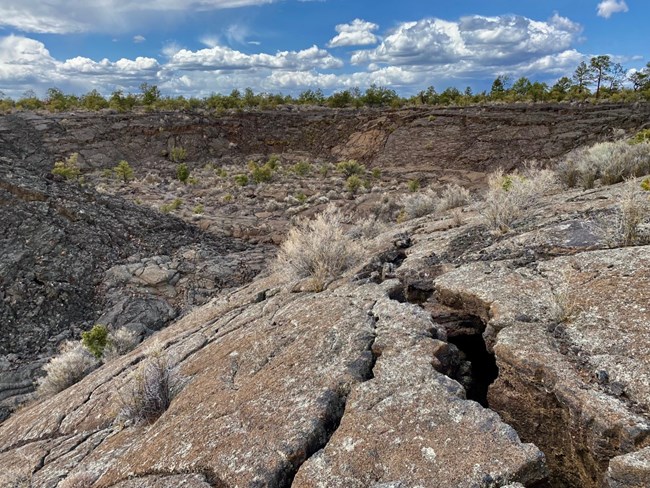 A circular ridge of smooth, cracked lava surrounds a depression. There are trees growing on the lava rock in the distance beyond the depression. Blue sky and puffy clouds are in the background.