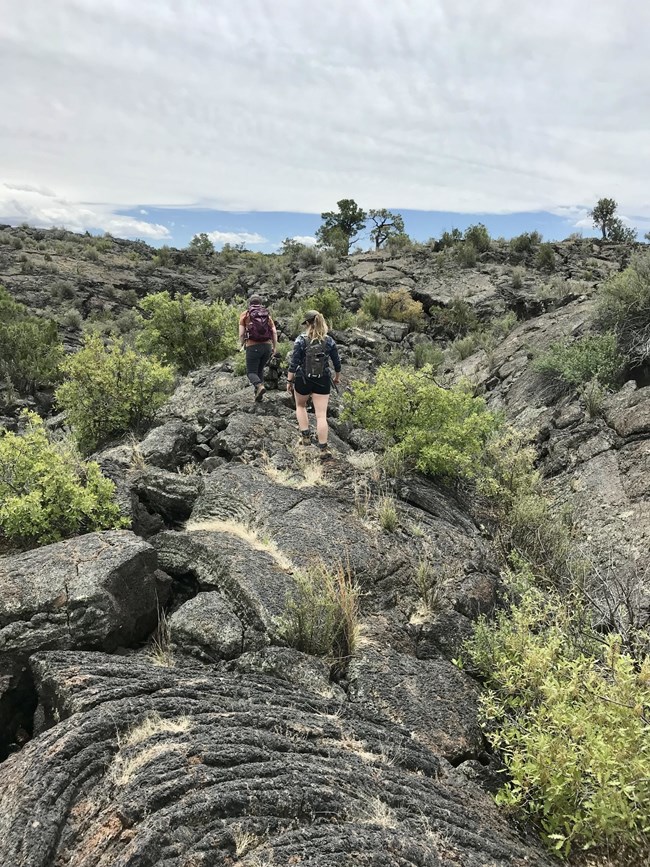 Two women are hiking on rugged lava. There is a cloudy sky in the background.