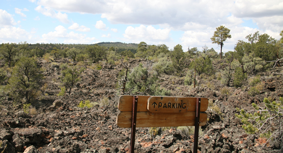A wooden sign with an upward-facing arrow reads "Parking."  It is the only man-made feature on a rugged lava flow dotted with bushes.