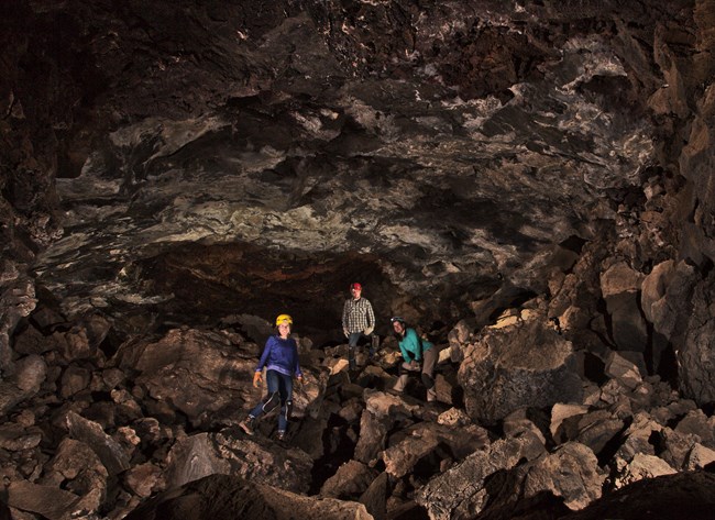 Three cavers stand on a floor of boulders beneath a craggy lava tube ceiling.