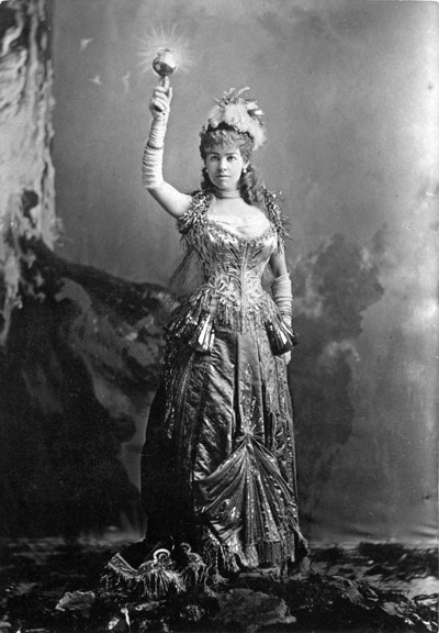 A woman dressed in an ornate, metallic ballgown. She is also wearing long gloves and feathered hat. One of her arms is raised and in that hand she is holding a torch.