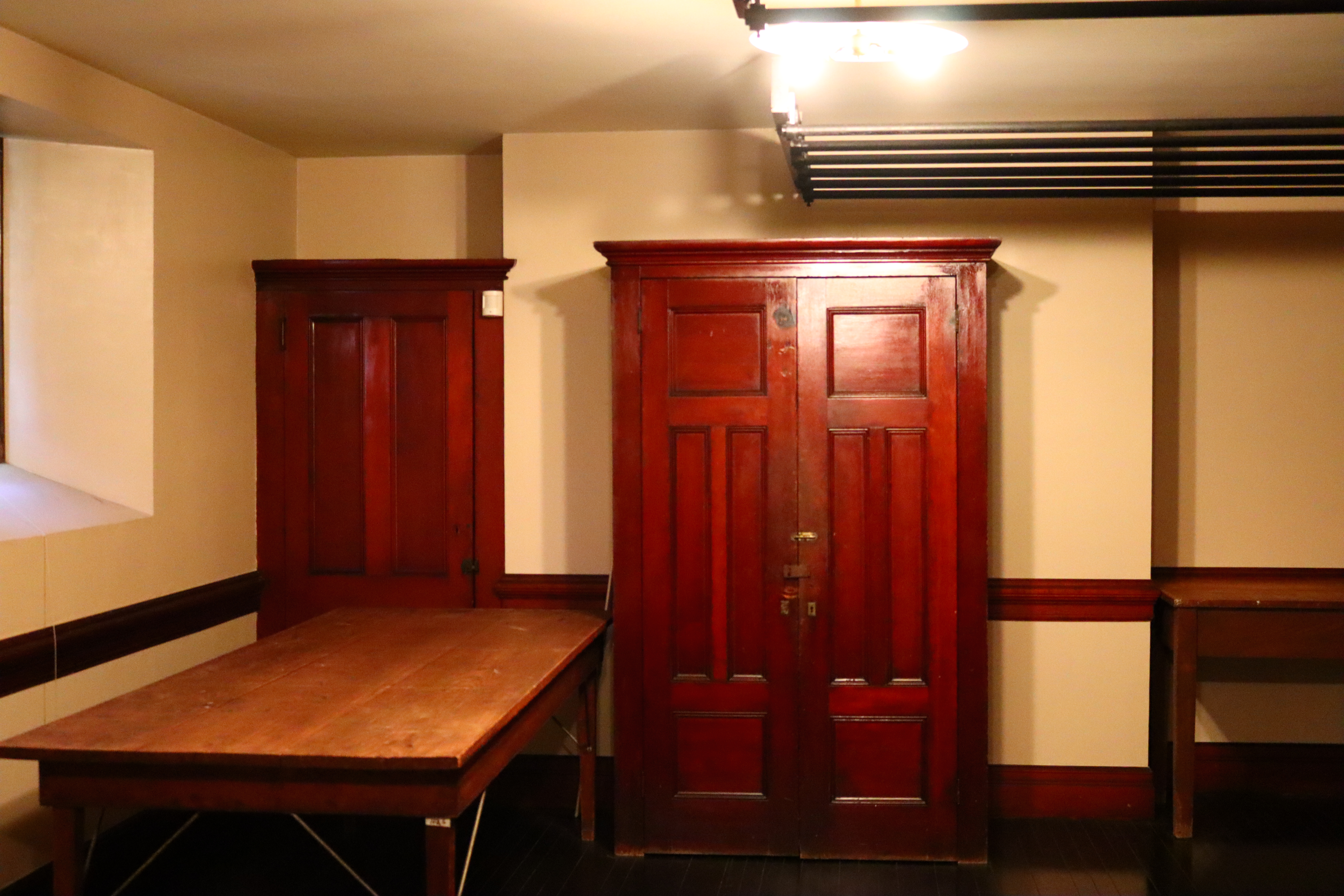 A tall wooden cabinet in a room with white walls. There is a table and a second cabinet to the left.