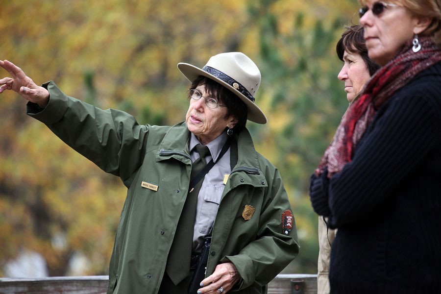 A woman wearing a Stetson-style hat and a green coat and tie points to something out of frame. Two other women stand to her right.