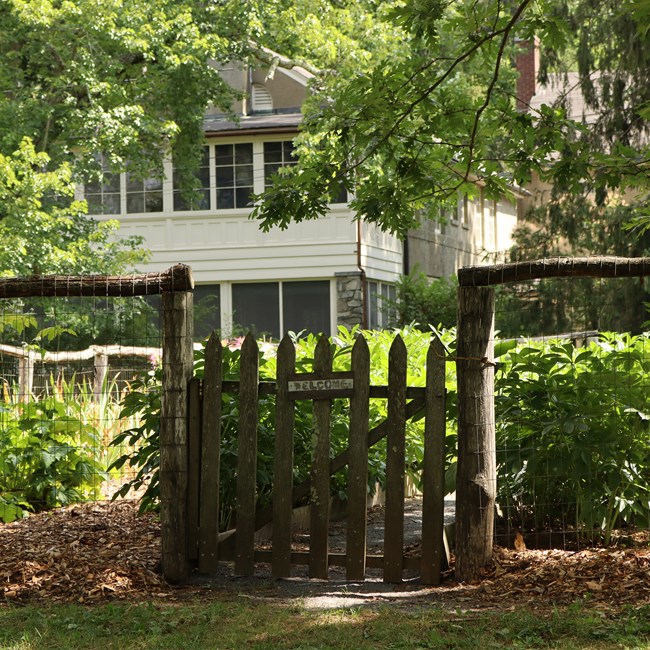 A wooden gate in front of a green garden. A small wooden plaque on the gate reads, "Welcome". In the background, a two-story cottage peaks through the trees.