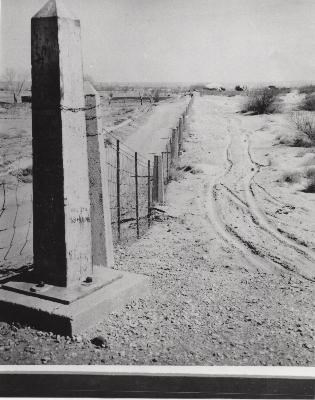 Una foto en blanco y negro de un monumento en forma de obelisco a la cabeza de una línea de postes de hormigón con la misma forma, conectados con malla de alambre.