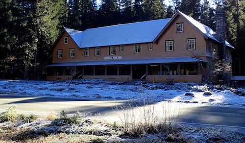 Un edificio de madera con tejados a dos aguas en sus extremos y un porche cubierto a su alrededor, bajo una capa de nieve y rodeado por un bosque.