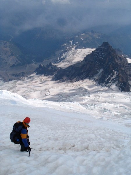 Un alpinista en una empinada ladera cubierta de nieve con vistas a una cumbre rocosa a menor altura y a valles distantes.