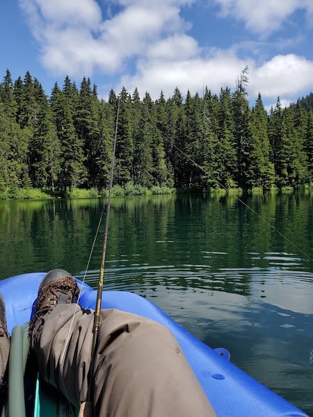 Imagen propia de una persona pescando desde una embarcación, con la caña apoyada en su rodilla, desde un bote flotando en un tranquilo lago circundado por un bosque.