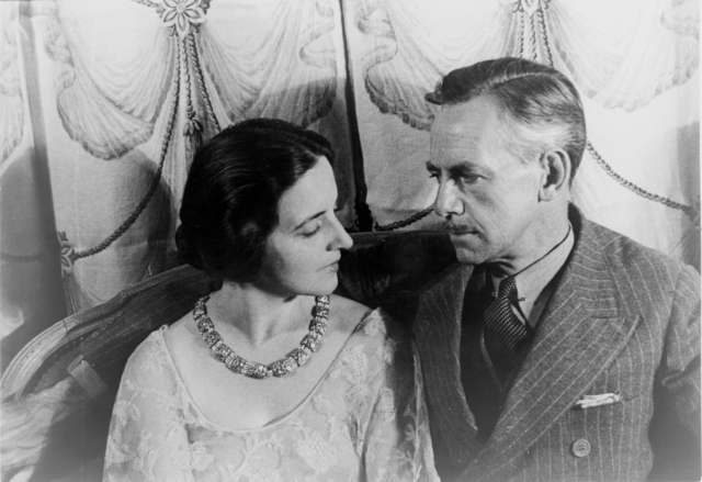 A black and white portrait of Carlotta Monterey and Eugene O'Neill. Carlotta, seated on the left, is wearing an elegant dress with a floral necklace. She has short, dark hair styled neatly and is looking intently at Eugene O'Neill.