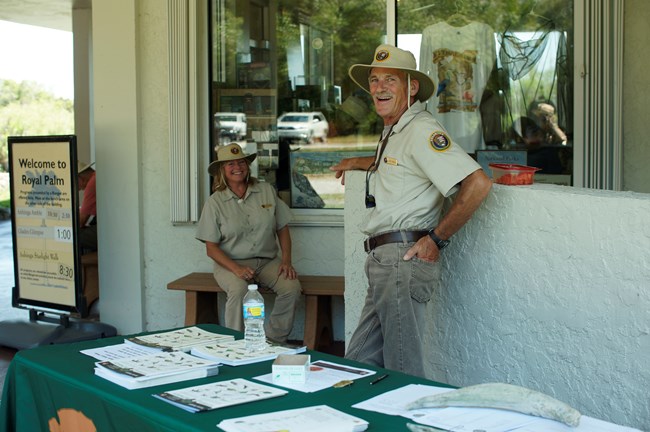 In the shade of a structure, a man in a volunteer uniform leans against a wall smiling at the camera while a woman in a volunteer uniform also is smiling at the camera while sitting down.