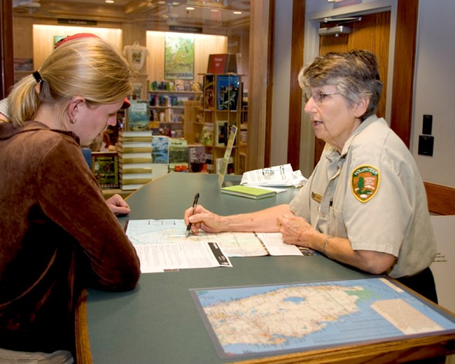 At a standing desk, a woman visitor leans on the desk as a volunteer assists her with orientation to the park while looking and pointing at a map.