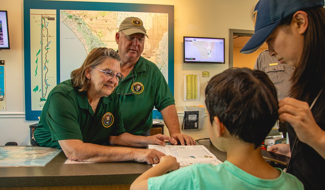 Two Park Volunteers behind a desk at the Visitor Center point to papers while helping a kid and their guardian.