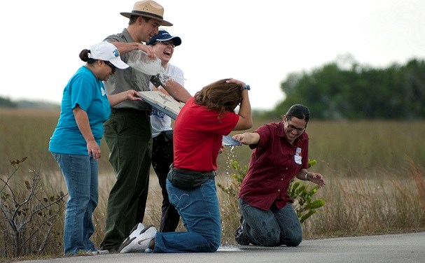park ranger pours water on board while adults standing and kneeling laugh