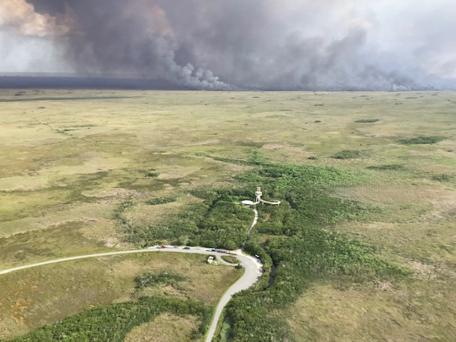 An aerial view shows a fire burning in the distance with a road and observation tower towards the front.