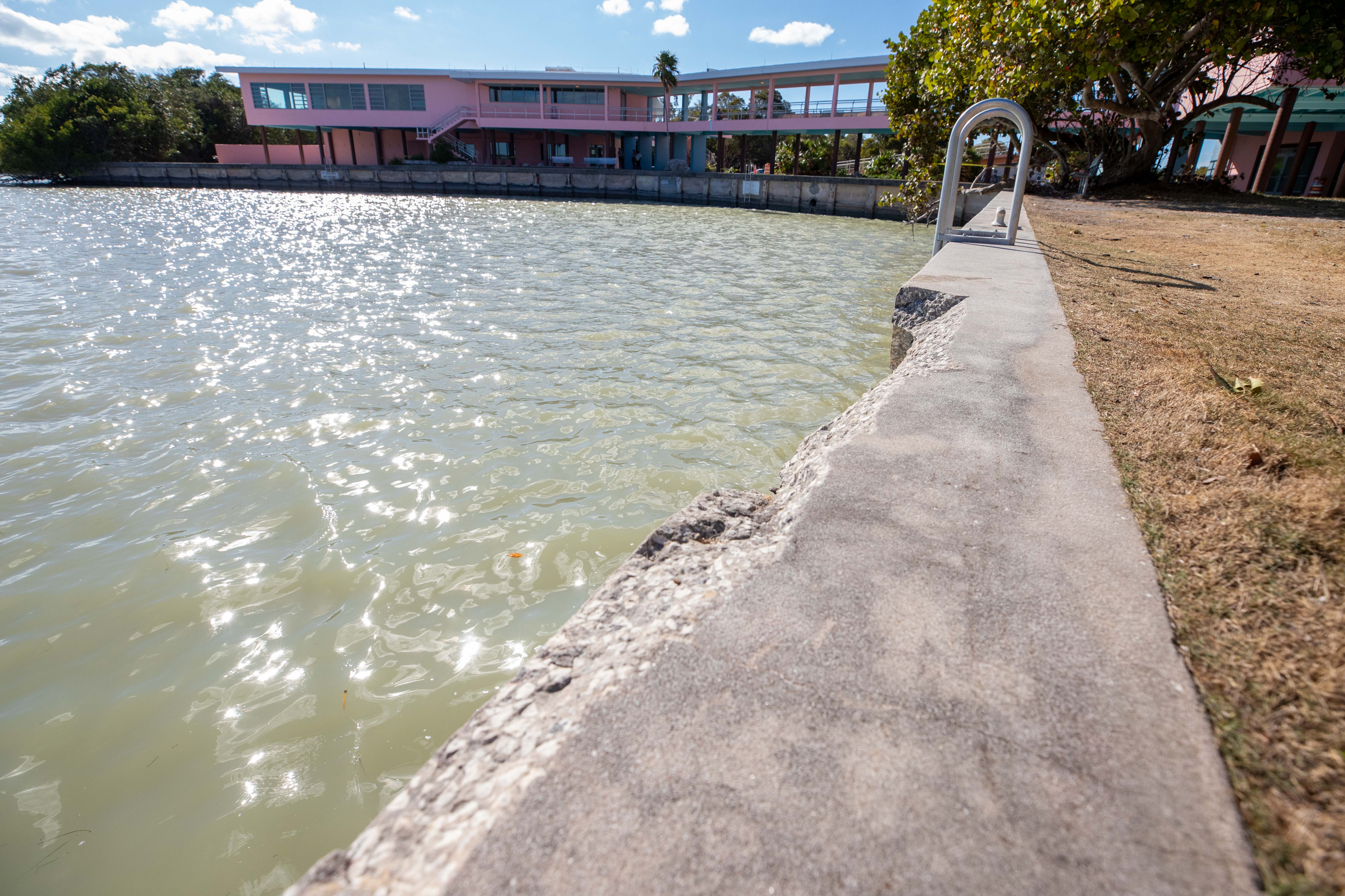 A cement seawall with a longitudinal section missing lines a boat basin with the two-story pink building with open breezeway in the background.