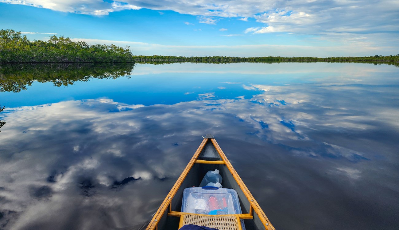 A yellow canoe moves through water. A cloudy blue sky and islands filled with mangroves and other vegetation are reflected on a body of water.