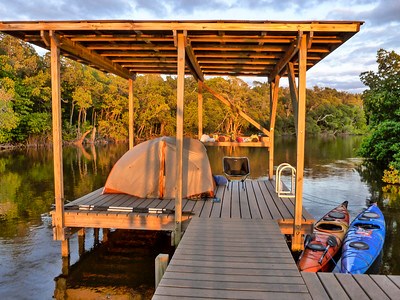 A tent on a lifted, wooden platform that sits above water. 2 kayaks are tied to the side, next to trees.