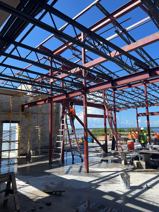 Inside a building under construction with steel beam framework, man on a ladder, and view of open water and mangroves in distance