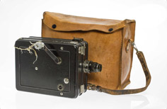 Old camera with black, rectangle-shaped casing next to brown camera bag.