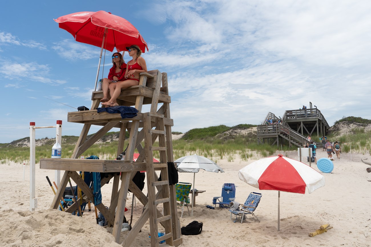 Two female lifeguards sit high in a chair on a sandy beach.