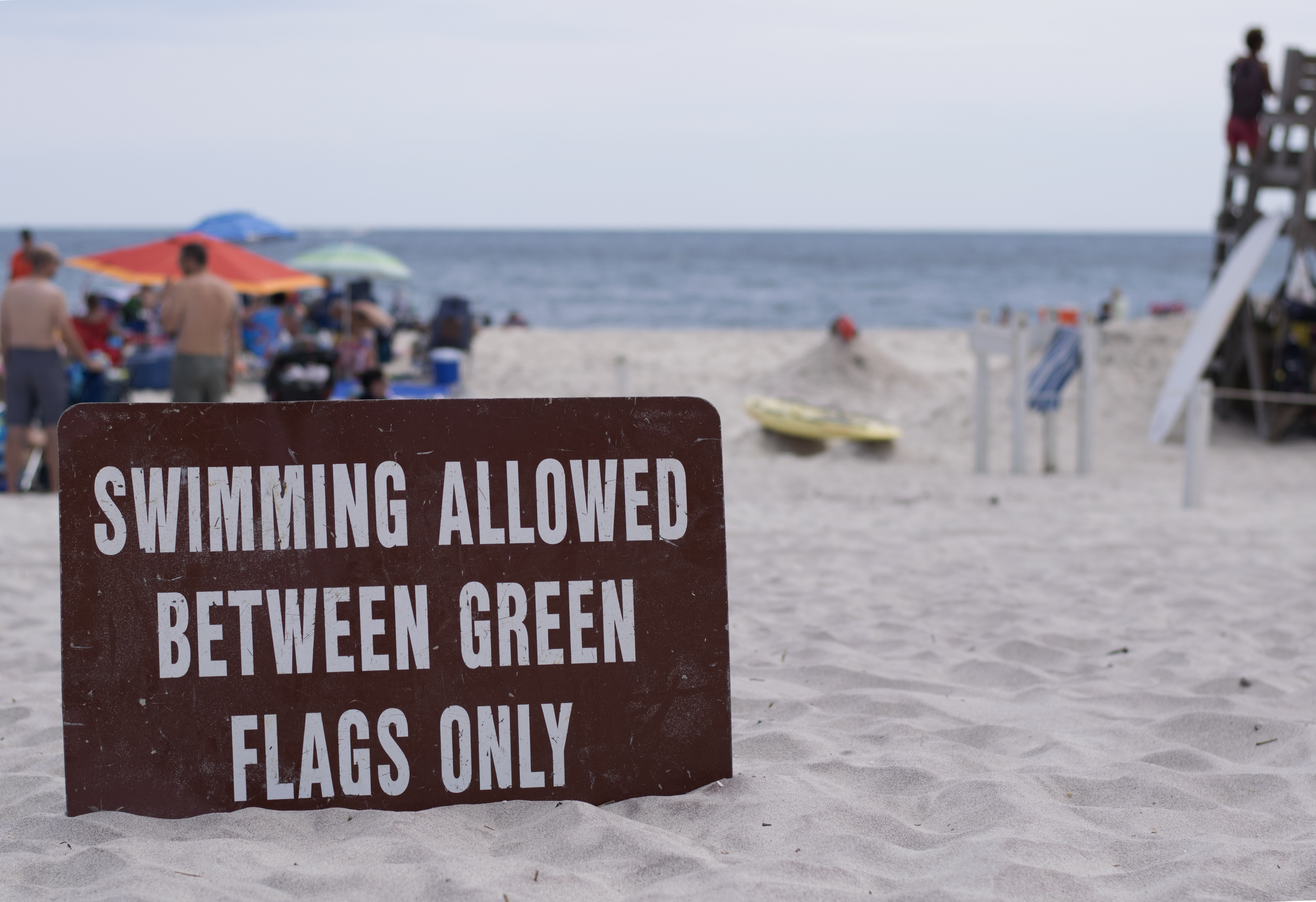 A sign reading, "Swimming allowed between green flags only" sits in the sand near a lifeguarded beach.