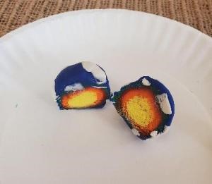 A play dough model of Earth cut in half sits on a paper plate. The inside is exposed showing a yellow center with layers of orange, red, green, and blue with bits of white encircling each other.