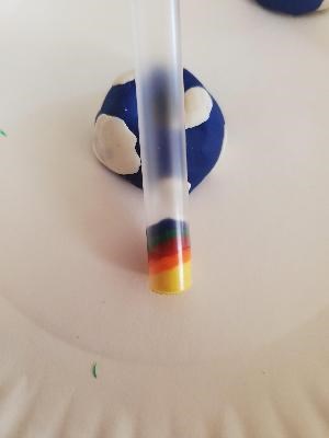 A clear straw rests against half of a play dough model earth. Layers of yellow, orange, red, green, and blue are stuck inside the straw.