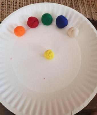 Six balls of play dough are arranged on a paper plate colored yellow, orange, red, green, blue, and white.