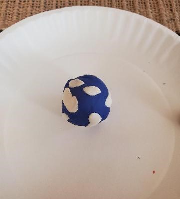 A play dough model of Earth showing the outermost layer of blue with bits of white for clouds sits on a paper plate.