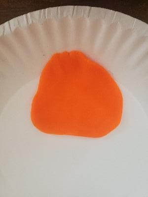 Orange play dough flattened into an irregular round shape on a paper plate.