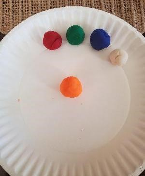 An orange play dough ball is in the center of a paper plate with smaller red, green, blue, and white balls behind it.