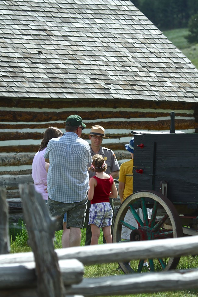 A group of people stand in front of a historic cabin. There is a historic wagon next to them.