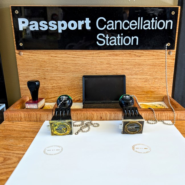 A sign that reads passport cancellation station sits over a small wooden desk with two rubber stamps and a piece of paper.