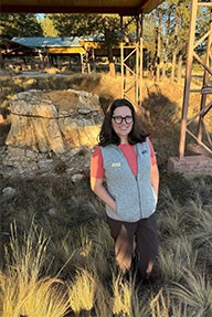 Woman with brown hair, wearing a pink shirt and glasses is standing in front of a fossilized tree stump.