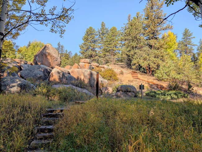 Stone stairs climb a small hill toward granite boulders. Wooden stairs climb the hillside behind the boulders.