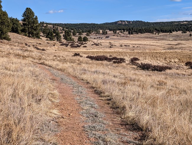 A red gavel trail runs along the right side of the photo. A tan grassy valley takes up most of the photo with a historic house and outbuildings in the background.