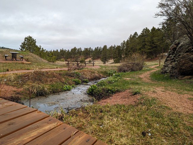 A bridge leads over a stream to several picnic tables. A trail winds past a rock formation on the right toward a hillside covered in pine trees.