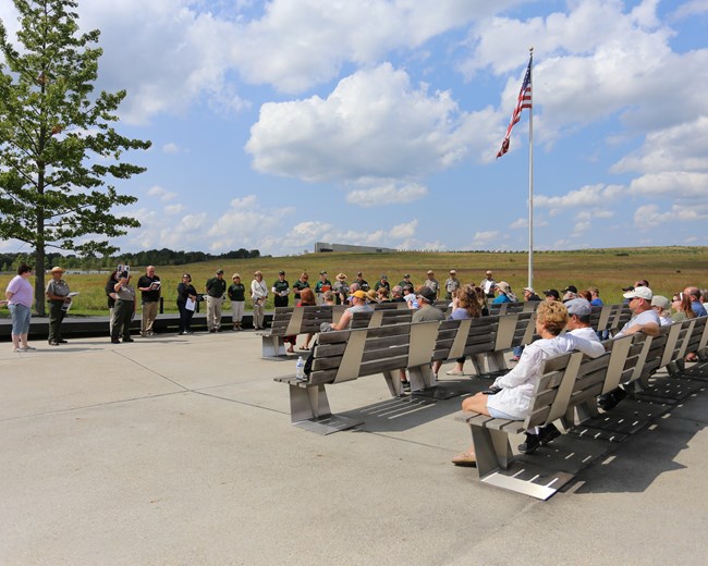 Visitors sitting outside on benches listening to rangers give a program next to large flag pole with American Flag on a sunny day with blue skies overhead.