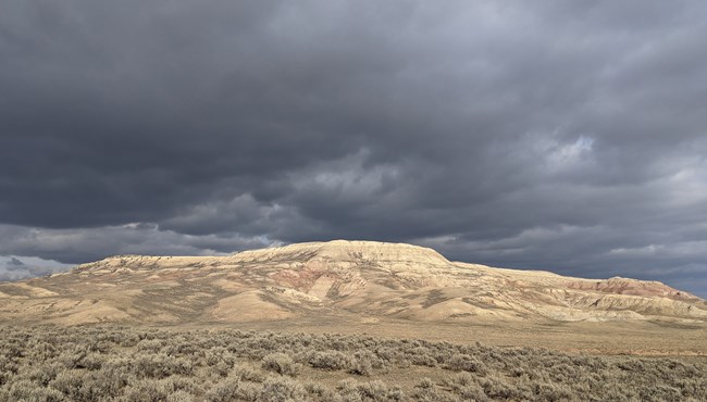 Dark gray clouds above a tan butte with sagebrush below.