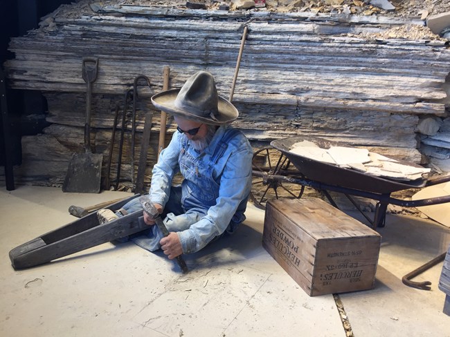 A mannequin with a peg leg uses a hammer and chisel on rock.