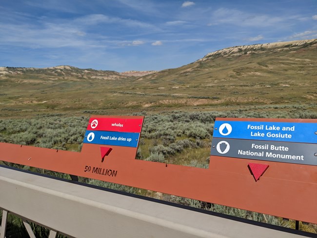 A railing with labels above the words 50 million for "whales" and "Fossil Lake dries up" and labels for "Fossil Lake and Lake Gosiute" and "Fossil Butte National Monument" to the right. Part of Fossil Butte and another ridge are visible behind.