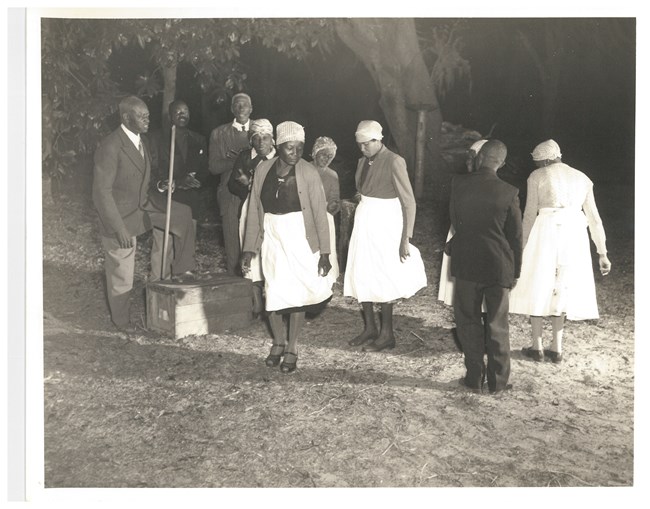 Black and white photograph of African Americans performing a Ring Shout, three men stand in the background while a group of men and woman form a circle in the foreground.