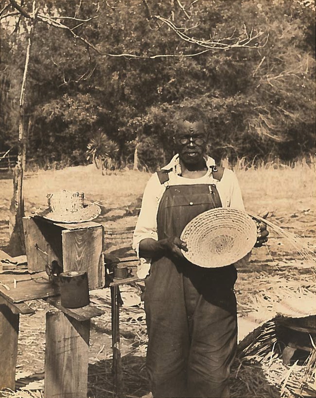 Charles Wilson, African American basket maker holding a sweetgrass basket and wearing a white shirt, overalls and glasses.