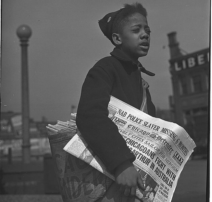 Young African American boy with coat and hat on displaying Chicago Defender newspaper and a satchel full pf papers slung over his shoulder.