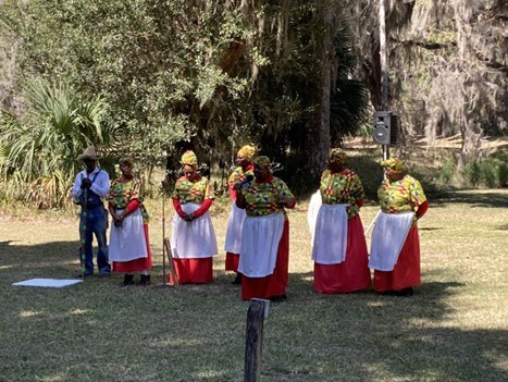 Gullah Geechee ring shouters in vibrant red, green and yellow waiting to perform with a speaker holding a microphone at center.