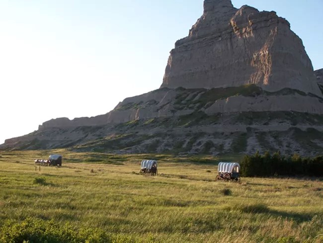 A large, rugged rock formation rises prominently against a clear blue sky, showcasing its natural textures and colors, with covered wagons traveling past it.