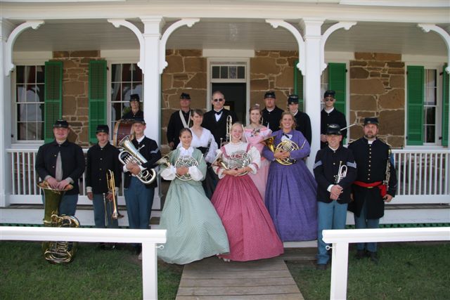 A color photograph of a group of people dressed in 19th century uniforms and clothing pose with instruments on the porch of a sandstone building.